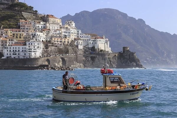 Fisherman in fishing boat heads out to sea from Amalfi harbour, with view towards Amalfi town
