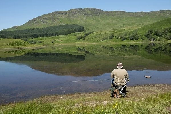 Angler sitting on shore of freshwater loch, Loch Lubhair, near Crianlarich, Trossachs, Stirling, Scotland, July