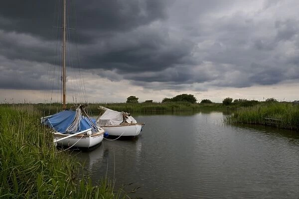 Boats moored under stormclouds, River Thurne, The Broads N. P. Norfolk, England, may
