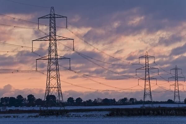 Electricity transmission pylons and overhead wires, crossing over snow covered farmland at sunset, Dorset, England