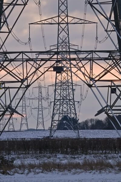 Electricity transmission pylons and overhead wires, crossing over snow covered farmland at sunset, Dorset, England, december