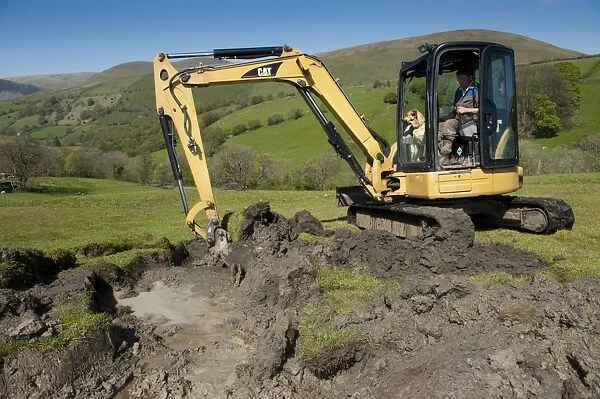 Farmer repairing old blocked drain in upland meadow using mini digger, Cumbria, England, May