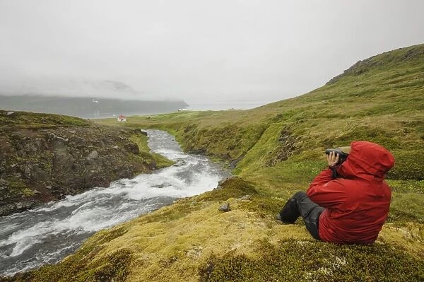 Hiker using binoculars, sitting beside river and coastline with low cloud, Hornstrandir, Westfjords, Iceland, July
