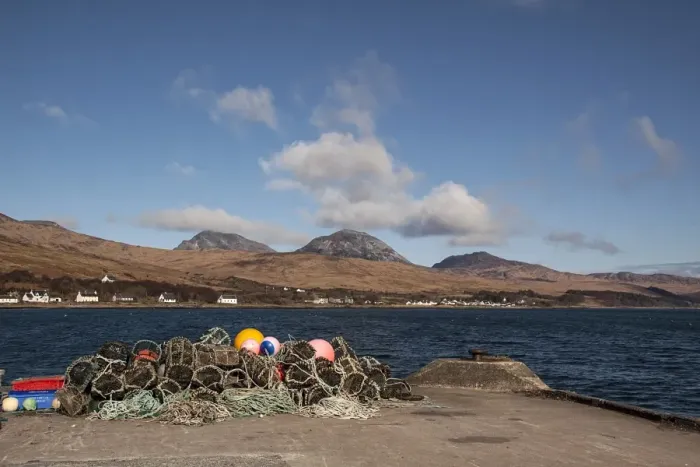 Framed Print : Lobster pots on Jura Pier with the village of Craighouse and the Paps of Jura Framed Print : Lobster pots on Jura Pier with the village of Craighouse and the Paps of Jura