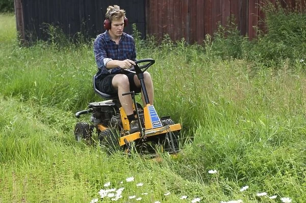 Man using ride-on mower on farm, cutting long grass around sheds, Sweden