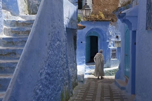 Man wearing djellaba walking beside blue buildings and stairway in alley of city, Chefchaouen, Morocco, april