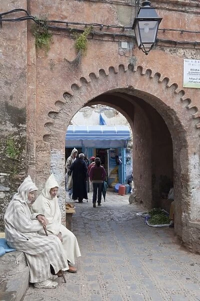 Men wearing djellaba sitting beside medina archway in city, Chefchaouen, Morocco, april