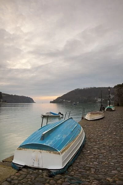 Small dinghies laying upside down on cobbled quay, with overcast sky at sunrise, Dartmouth Harbour, Dartmouth, Devon