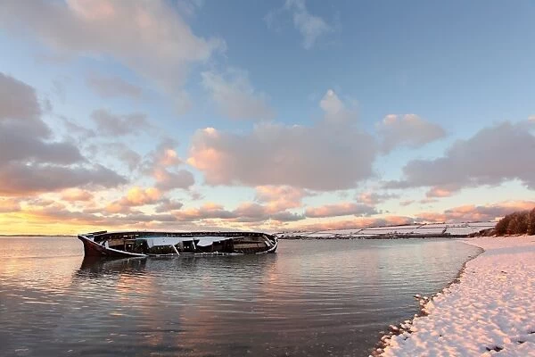 Snow covered riverbank and wreck of old barge partly submerged by incoming tidal river at sunset, River Taw