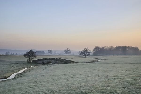 View of arched bridge over beck flowing through parkland at dawn, Hovingham Park, Hovingham, North Yorkshire, England