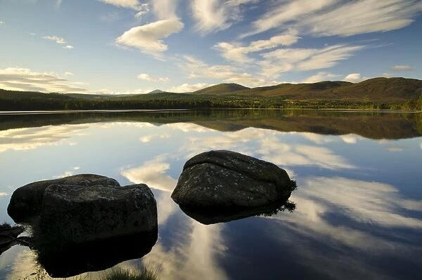 View of blue sky, clouds and rocks reflected in still water of freshwater loch, Loch Garten, Abernethy Forest