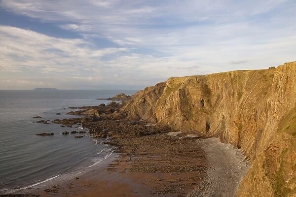 View of coastal cliff with folded strata in late afternoon sunlight, with Lundy Island on horizon in distance
