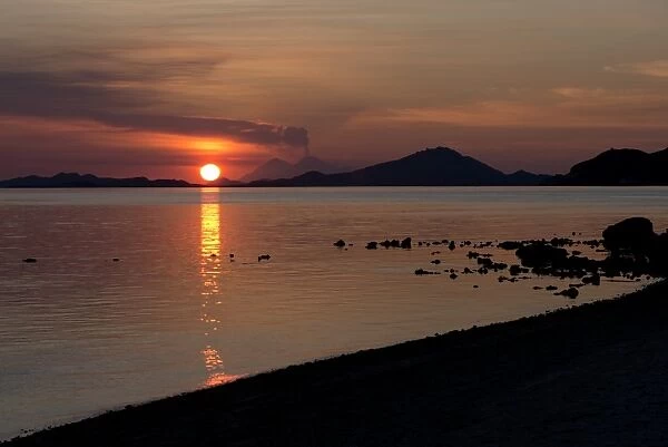 View of coastline with erupting volcano in distance at sunset, Sangeang Api (Mount Sangeang), Kelor Island