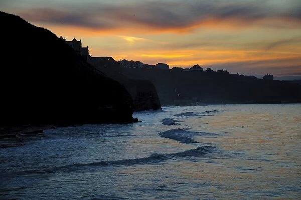 View of sea cliffs and clifftop seaside town silhouetted at sunset, West Cliff, Whitby, North Yorkshire, England, March