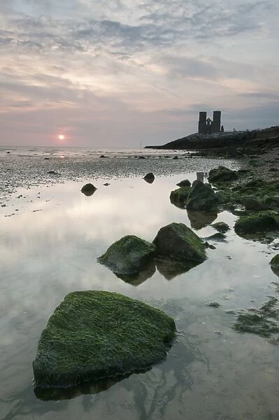 View of seaweed covered rocks beach at low tide, with 12th Century ruined church in distance at sunrise, St
