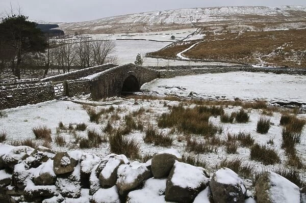 View of stream flowing through snow covered fields and under stone bridge, with ridge of Widdale Fell in background