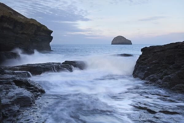View of waves crashing through manmade gully in coastal rocks at sunrise, with Gull Rock just off headland at Dennis