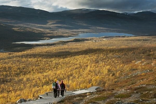 Walkers on boardwalk leading to summit of fell, Malla Strict Nature Reserve and Lake Kilpisjarvi in background