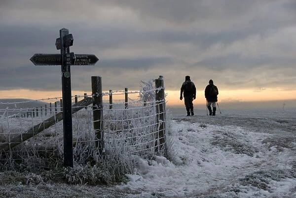 Walkers on frost and snow covered footpath, beside signpost and fence at sunset, Ridgeway Path, near Pitstone Hill, Chilterns, Buckinghamshire, England, december