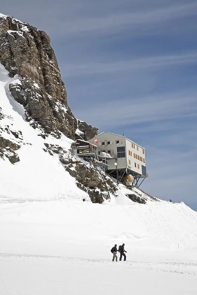 Walkers walking on snow covered mountain below mountain hut, Monchsjoch Hut, Jungfraujoch, Bernese Alps, Switzerland