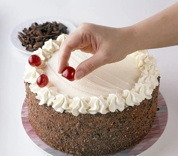 Black forest gateau, cherries being placed around the edges, plate of chocolate shavings in the background, close-up