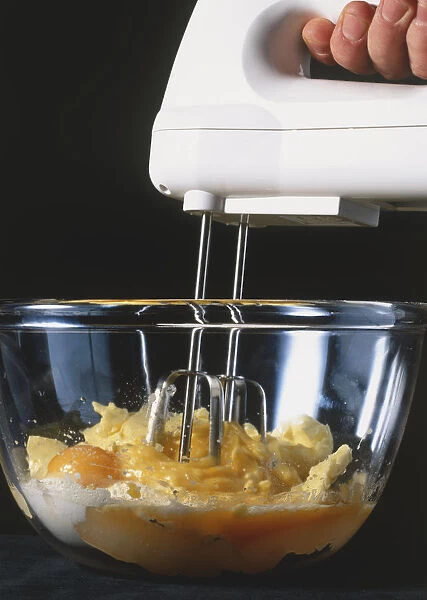 Hand-held electrical food mixer whisking eggs, flour and butter in glass bowl, side view