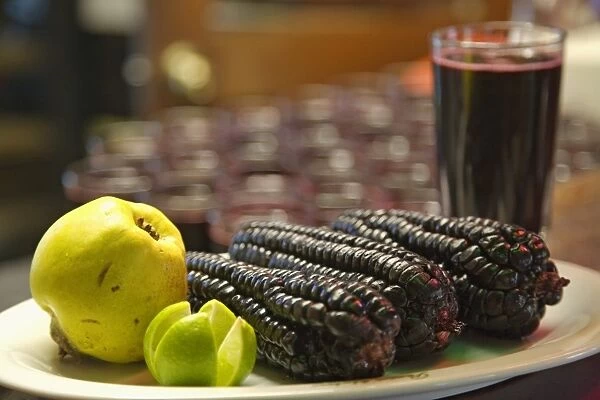 Peru, Lima, three purple maize corn cobs, a quince, and a sliced lime on a plate plate, glass of chicha morada in the background
