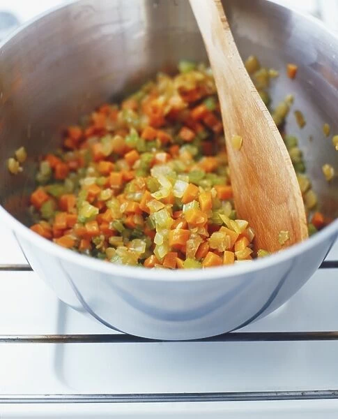 Using a wooden spoon to stir mixture of diced carrots, onion and celery cooking in a pan, high angle view