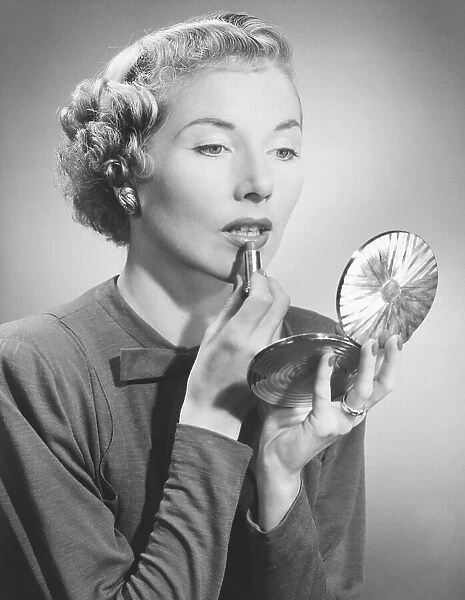 Woman looking at hand mirror, applying lipstick, (B&W)