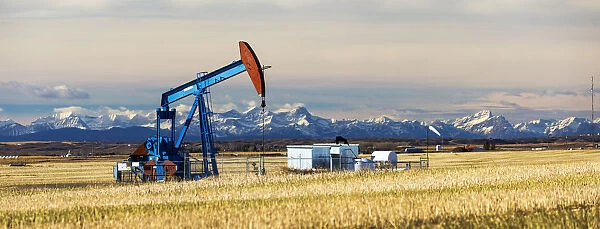 Panorama Of A Colourful Pump Jack In Cut Canola Stubble Field With Snow Capped Mountains, Blue Sky And Clouds In The Background; Alberta, Canada