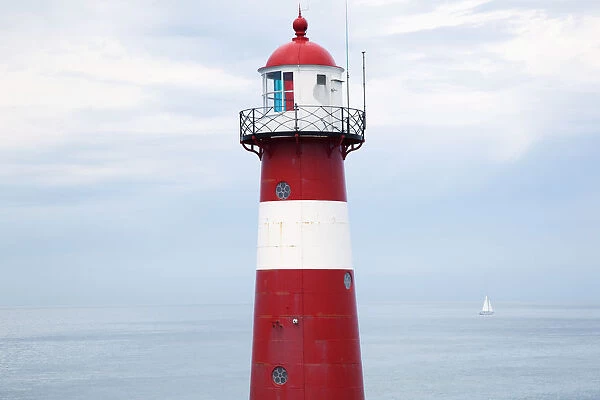 Red and white lighthouse along the coast with a sailboat in the distance near westkapelle; Zealand, netherlands