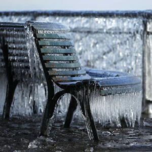 A frozen public bench is seen next to a lake side due to the heavy wind conditions