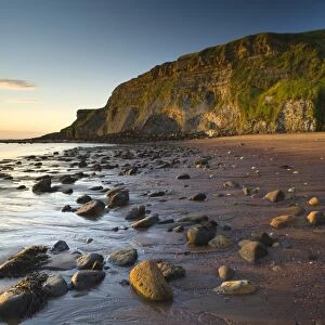 Rocks on sandy beach with sea cliffs at sunrise, Black Nab, Saltwick Bay, North Yorkshire, England, august