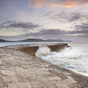 View of harbour wall with waves breaking at sunrise, looking across bay to Charmouth, Stonebarrow