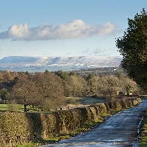 View of icy rural road, hedgerow and bare trees towards snow covered hill in distance, looking from Little Bowland Road toward Pendle Hill, Chipping, Lancashire, England, december