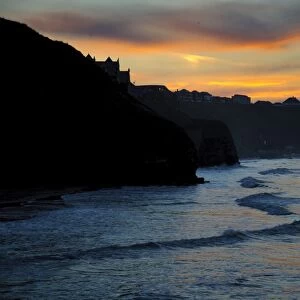 View of sea cliffs and clifftop seaside town silhouetted at sunset, West Cliff, Whitby, North Yorkshire, England, March