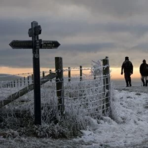 Walkers on frost and snow covered footpath, beside signpost and fence at sunset, Ridgeway Path, near Pitstone Hill, Chilterns, Buckinghamshire, England, december