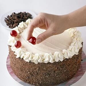 Black forest gateau, cherries being placed around the edges, plate of chocolate shavings in the background, close-up
