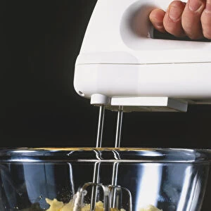 Hand-held electrical food mixer whisking eggs, flour and butter in glass bowl, side view