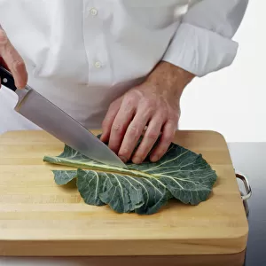 Using a chefs knife to cut leaf of spring greens on either side of the rib on wooden chopping board