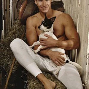 Man with a naked torso wearing long underwear with a dog in a horse barn sitting on bales of hay