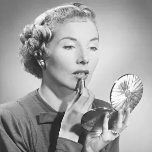 Woman looking at hand mirror, applying lipstick, (B&W)