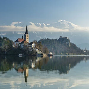 Looking across the calm waters of Lake Bled at dawn to the church on Lake Bled Island and Bled Castle behind; Bled, Slovenia