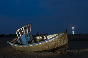 Shingle Prints: Abandoned fishing boat on shingle beach with distant lighthouse at night