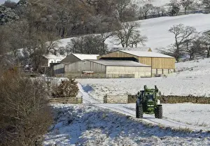 John Deere tractor travelling along snow covered farm track, with farm buildings in background, Whitewell, Lancashire, England, november