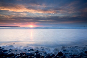 Pebbles Prints: View of beach with pebble ridge as wave receeds back into sea at sunset